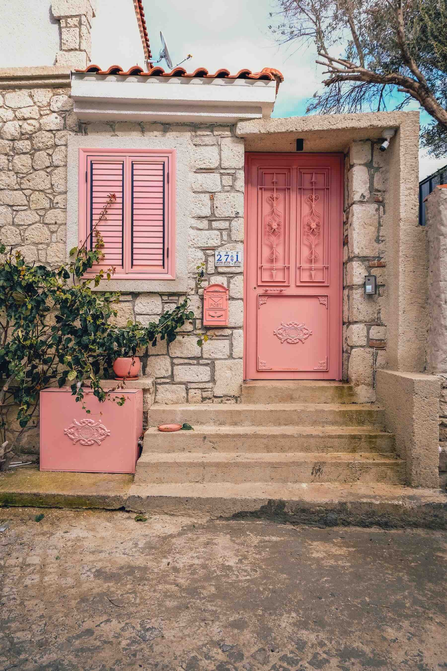 Exterior of a small stone cottage in Alaçatı, Turkey with a light pink front door and matching window shutters.
