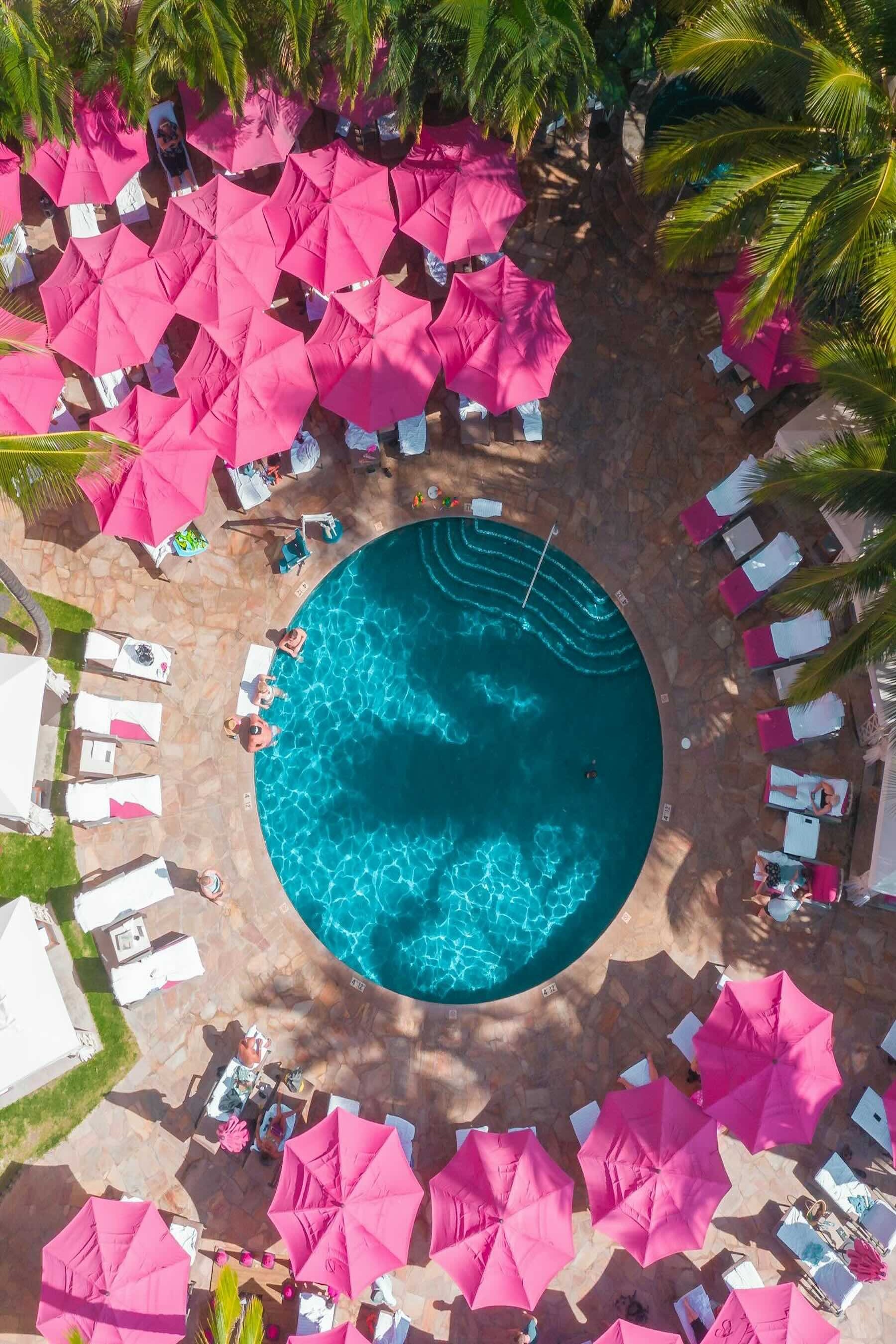Birdseye view of the circular hotel pool at the Royal Hawaiian, Waikiki. The pool is surrounded by sunloungers, large fuchsia pink sun umberallas and palm trees.